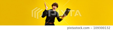 Banner. Portrait of young attractive man shouting of joy with laptop against vivid yellow studio background with negative space for text. Banner. Portrait of young attractive man shouting of joy with laptop against vivid yellow studio background with negative space for text. 109300132