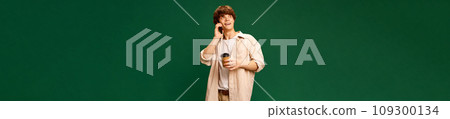 Banner. Young attractive ma student talking on phone and drinking coffee against green studio background with negative space for text. Banner. Young attractive ma student talking on phone and drinking coffee against green studio background with negative space for text. 109300134