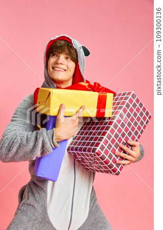Portrait of charming young man standing and thinking about who should be first to give big gift from those that he holds against pink background Portrait of charming young man standing and thinking about who should be first to give big gift from those that he holds against pink background 109300136