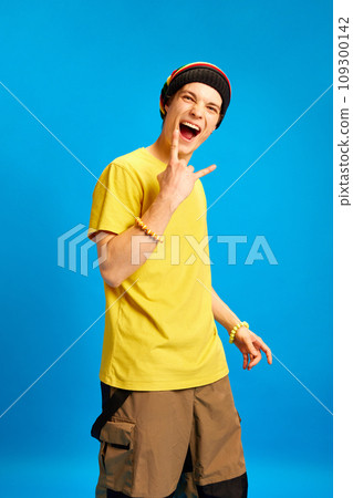Happy shouting young man wearing rastaman, reggae style outfit showing rock sign against blue studio background. Happy shouting young man wearing rastaman, reggae style outfit showing rock sign against blue studio background. 109300142