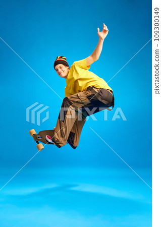 Positive, smiling man wearing in rastaman, reggie style doing Ollie trick in motion against blue background. Positive, smiling man wearing in rastaman, reggie style doing Ollie trick in motion against blue background. 109300149