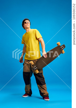 Positive, smiling man in sunglasses wearing in rastaman, reggie style and posing with skateboard against blue background. 109300152