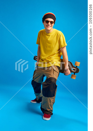 Full-length portrait of attractive cool attitude in reggae, rasta man style with skateboard against blue studio background. 109300166