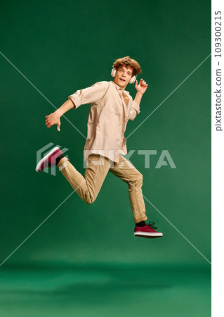 Full length portrait of young man, student dressed retro fashion outfit in headphones jumps high against green studio background. 109300215