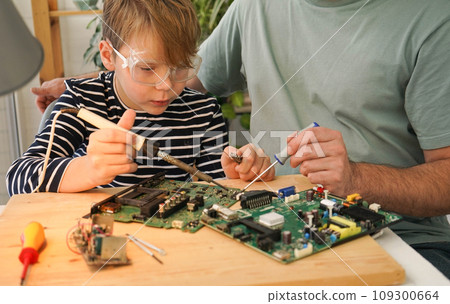 Father with his son spend time together. Close-up of a man teaching his boy at home to solder computer spare parts. Education moment during parenthood. Togetherness concept. 109300664