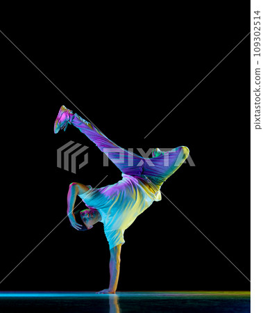 Breakdancer. Young flexible athletic man dancing breakdance freezes in sport outfit against black background in neon light. 109302514