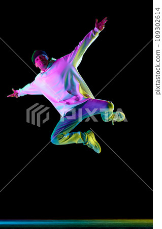 Young male dancer, athletic guy dancing freestyle, hip-hop tricks in motion in stylish outfit against black background in neon light. 109302614