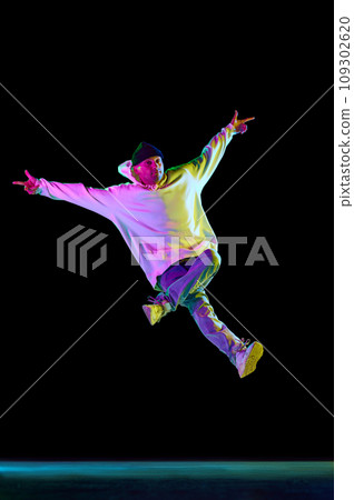 Studio shot of young flexible sportive man dancing breakdance, freezing elements in casual outfit against black background in neon light Studio shot of young flexible sportive man dancing breakdance, freezing elements in casual outfit against black background in neon light 109302620