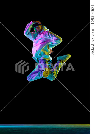 Studio shot of young flexible sportive man dancing breakdance, jumping, freezing in casual outfit against black background in neon light Studio shot of young flexible sportive man dancing breakdance, jumping, freezing in casual outfit against black background in neon light 109302621