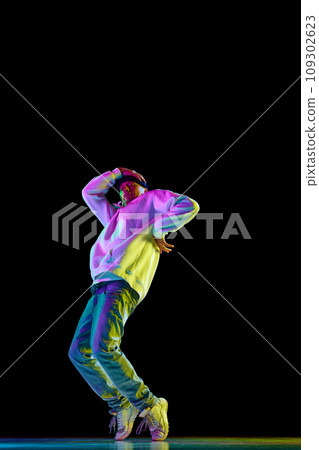 Young stylish man, break dancing dancer practicing solo in casual outfit against black background in neon pink light at dance hall. Young stylish man, break dancing dancer practicing solo in casual outfit against black background in neon pink light at dance hall. 109302623