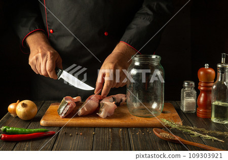 Cutting fish into steaks before salting in a jar. The concept of preparing a fish dish or herring with the hands of a chef in the kitchen Cutting fish into steaks before salting in a jar. The concept of preparing a fish dish or herring with the hands of a chef in the kitchen 109303921