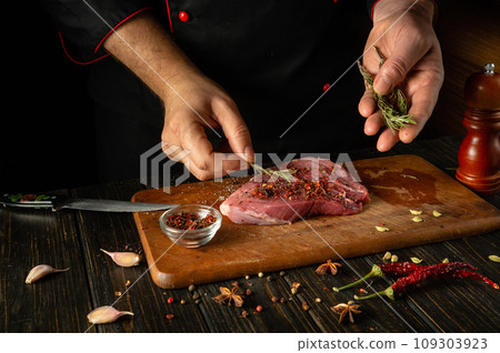 The chef adds dry aromatic rosemary to the meat steak. The process of cooking veal meat or shish kebab on the kitchen table with spices and salt 109303923