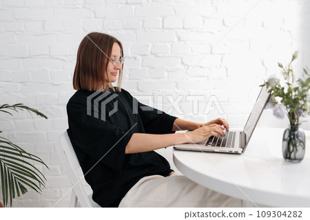 Smiling young woman in glasses holds online meeting via laptop. Businesswoman, entrepreneur, freelancer works remote. Student studies online with teacher on skype, watches webinar, typing on keyboard 109304282