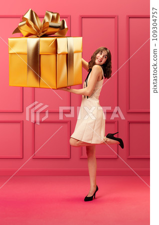 Pretty, elegant young girl holding giant golden present box, Christmas gift against pink background. Happiness, surprise 109304757