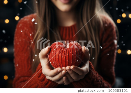 A young woman with long, curly hair, wearing a red sweater. She is holding a red, knitted Christmas heart in her hands. 109305101