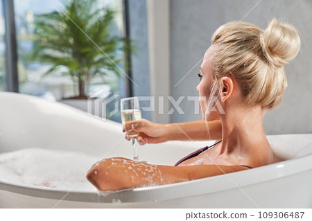Unrecognizable blonde woman taking glass of sparkling wine, while lying in bathtub. Back view of relaxed lady with hairstyle soaking in bubble bath, against blurred background. Relax and spa concept. Unrecognizable blonde woman taking glass of sparkling wine, while lying in bathtub. Back view of relaxed lady with hairstyle soaking in bubble bath, against blurred background. Relax and spa concept. 109306487
