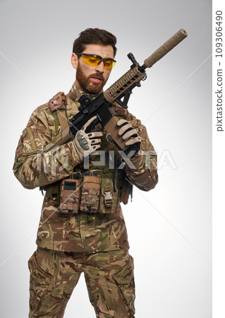 Focused male soldier in camouflage outfit recharging sniper rifle indoors. Portrait of caucasian military man in tactical gloves, looking at weapon, on gray background. War, military force concept. 109306490