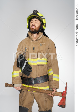 Exhausted caucasian firefighter with closed eyes, holding shabby axe. Front view of tired, bearded man in uniform with hatchet resting, on gray studio background. Concept of job, workplace, equipment. 109306500