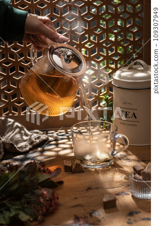 Tea is poured into a cup from a teapot with steam in bright sunlight shining into a warm atmosphere, wooden table Tea is poured into a cup from a teapot with steam in bright sunlight shining into a warm atmosphere, wooden table 109307949