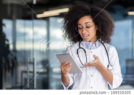 A young female doctor holds a tablet computer in her hands, a woman in a white medical coat consults patients remotely, smiles with satisfaction, stands by the window. A young female doctor holds a tablet computer in her hands, a woman in a white medical coat consults patients remotely, smiles with satisfaction, stands by the window. 109310105