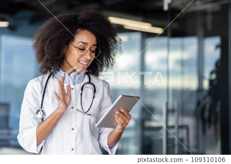 A young female doctor holds a tablet computer in her hands, a woman in a white medical coat consults patients remotely, smiles with satisfaction, stands by the window. A young female doctor holds a tablet computer in her hands, a woman in a white medical coat consults patients remotely, smiles with satisfaction, stands by the window. 109310106