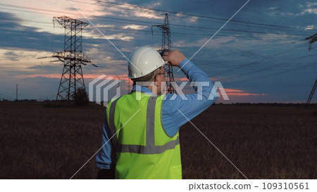 Engineer in helmet and safety vest past power transmission lines in semi dark field. Mature technician works night shift at power generation plant in country field. Electrician at power substation Engineer in helmet and safety vest past power transmission lines in semi dark field. Mature technician works night shift at power generation plant in country field. Electrician at power substation 109310561
