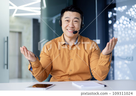 Portrait of a smiling young asian man sitting in the office at a table in front of a camera in a headset, spreading his hands, smiling at the camera. 109311024