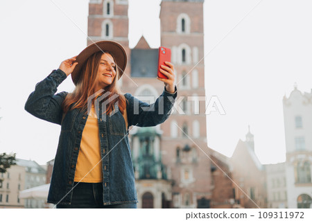 Young woman tourist in hat making selfie photo in front of the famous St. Mary's Basilica on the Market square in Krakow, Poland. Traveling Europe in autumn. Vacation concept Young woman tourist in hat making selfie photo in front of the famous St. Mary's Basilica on the Market square in Krakow, Poland. Traveling Europe in autumn. Vacation concept 109311972