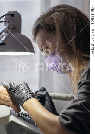 Manicurist at work. Professional manicurist removes old worn nail design from nails of client using modern electric drill. Preparing fingernails for making fresh manicure and applying new nail design 109316982