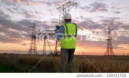 Engineer in helmet holds tablet walking against power transmission lines 109321726