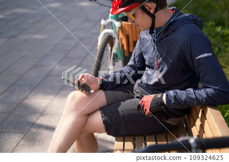 Man in a bicycle helmet sits on a bench and looks at his phone, resting while cycling in the city Man in a bicycle helmet sits on a bench and looks at his phone, resting while cycling in the city 109324625