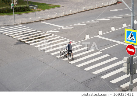 Man crosses the road at a pedestrian crossing and carries a bicycle next to him. View from above. Man crosses the road at a pedestrian crossing and carries a bicycle next to him. View from above. 109325431