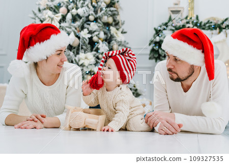 Happy family under Christmas tree. baby boy in Santa Claus hat with gifts under Christmas tree with many gift boxes presents. Happy Holidays, New year. Cozy warm winter evening at home. Xmas time 109327535