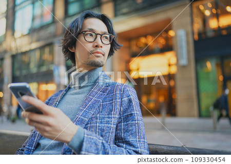 A man wearing a jacket sitting on a bench and using a smartphone A man wearing a jacket sitting on a bench and using a smartphone 109330454