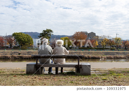 Friendly elderly couple sitting on a bench by the river Friendly elderly couple sitting on a bench by the river 109331975