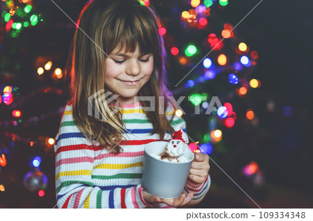 Little child girl holding cup with hot chocolate with marshmallows as Santa Claus. Kid by near Christmas tree decorated with lights. Little child girl holding cup with hot chocolate with marshmallows as Santa Claus. Kid by near Christmas tree decorated with lights. 109334348