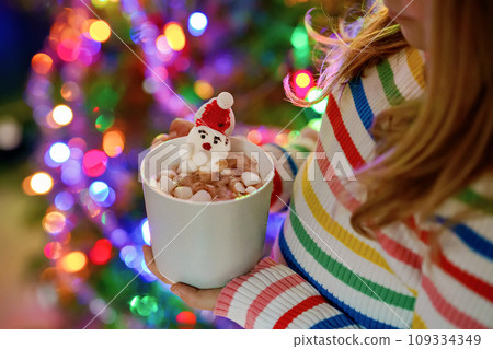 Little child girl holding cup with hot chocolate with marshmallows as Santa Claus. Kid by near Christmas tree decorated with lights. Little child girl holding cup with hot chocolate with marshmallows as Santa Claus. Kid by near Christmas tree decorated with lights. 109334349