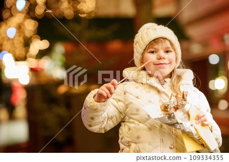 Little preschool girl, cute child eating churros sweets covered with chocolate with decoration and lights on background. Happy child on Christmas market in Germany. 109334355