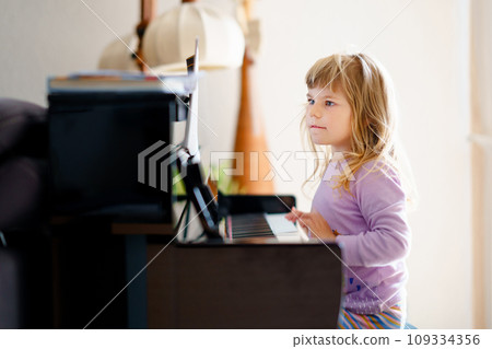 Beautiful little toddler girl playing piano in living room. Cute preschool child having fun with learning to play music instrument. Early musical education for children. Beautiful little toddler girl playing piano in living room. Cute preschool child having fun with learning to play music instrument. Early musical education for children. 109334356