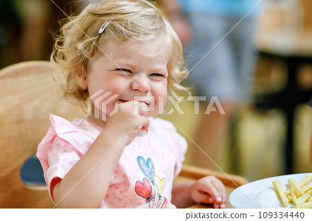 Adorable toddler girl eating healthy vegetables and unhealthy french fries potatoes. Cute happy baby child taking food from dish at daycare or nursery canteen. Adorable toddler girl eating healthy vegetables and unhealthy french fries potatoes. Cute happy baby child taking food from dish at daycare or nursery canteen. 109334440