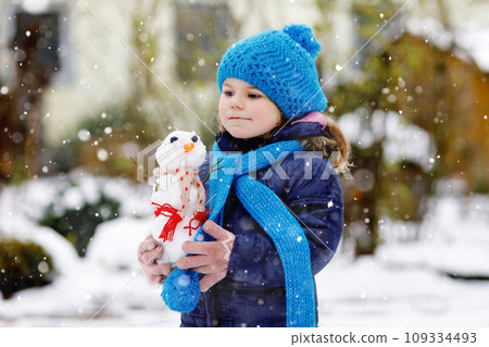 Cute little toddler girl making mini snowman and eating carrot nose. Adorable healthy happy child playing and having fun with snow, outdoors on cold day. Active leisure with children in winter 109334493