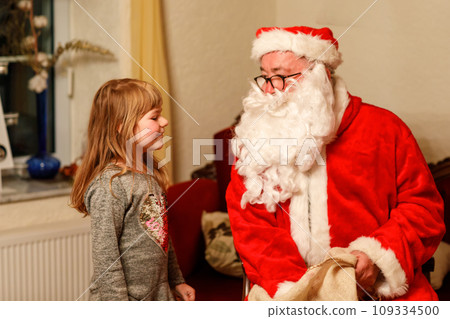 Little toddler girl talking to Santa Claus called Nikolaus or Weihnachtsmann in German. Happy smiling kid waiting for gifts. Santa with golden book. Xmas, childhood, holiday 109334500