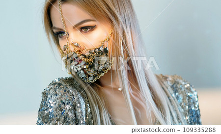 DIY mask accessory. Pandemic party. Blonde glamour woman in trend jewelry chain gemstone creative quarantine style matching outfit isolated on blue background. DIY mask accessory. Pandemic party. Blonde glamour woman in trend jewelry chain gemstone creative quarantine style matching outfit isolated on blue background. 109335288