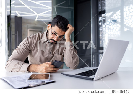 Upset sad man at workplace inside office, businessman uses phone reads bad news received by e-mail, employee disappointed holds smartphone in hands browses internet sites and social networks. 109336442