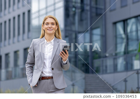 Portrait of a young businesswoman in a suit walking outside an office center, holding a phone and hand in her pocket, smiling and looking at the camera. 109336453