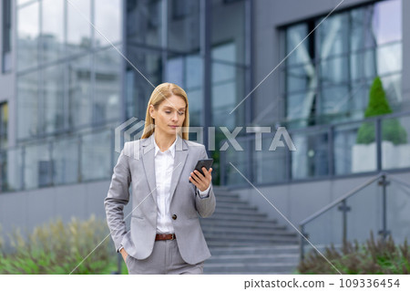 Young businesswoman in a suit standing near an office center and struggling with a mobile phone, writing a message, reading the news, chatting. 109336454