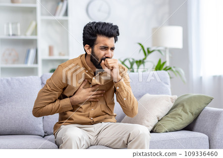 Indian young man sitting at home on the couch and coughing, covering his mouth, holding his chest, feeling severe pain, suffering from an infectious disease. 109336460