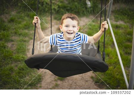 Little boy in striped t-shirt swinging in the swing-nest at a park on summer day Little boy in striped t-shirt swinging in the swing-nest at a park on summer day 109337202