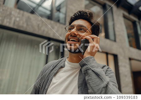 Smiling male manager in eyeglasses is talking phone standing in modern coworking space Smiling male manager in eyeglasses is talking phone standing in modern coworking space 109339183