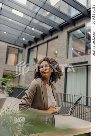 Smiling businesswoman holding laptop and looking at side while standing in modern coworking 109339188